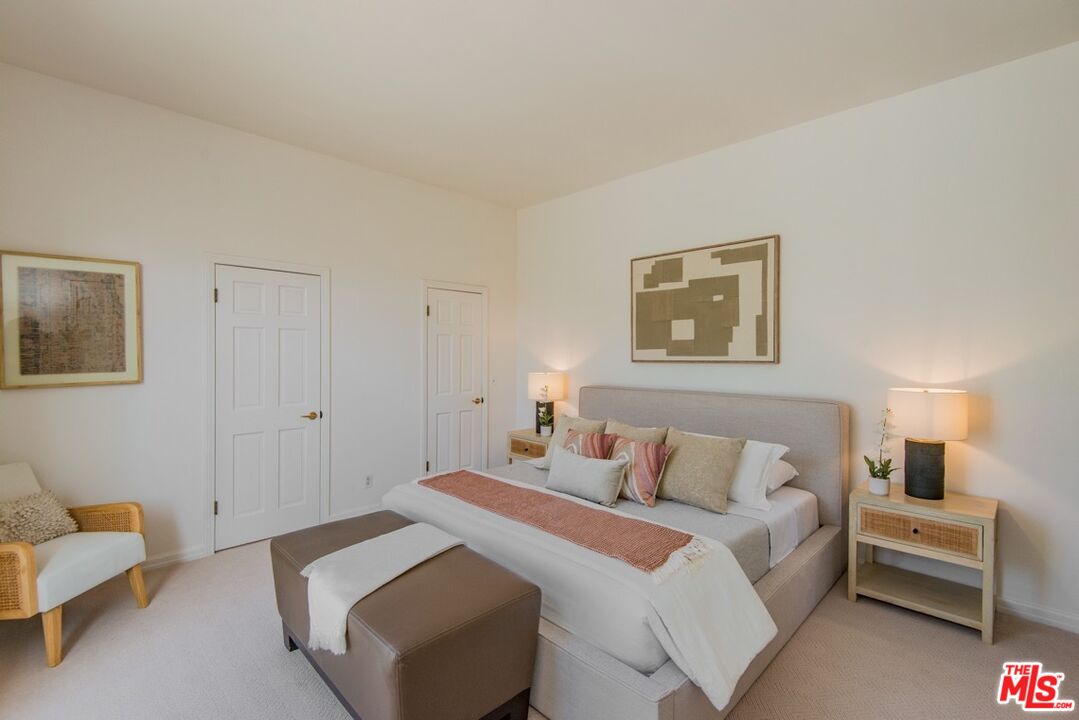 Staged bedroom with an upholstered bed, coral accent bedding, matching nightstands, and a brown ottoman.
