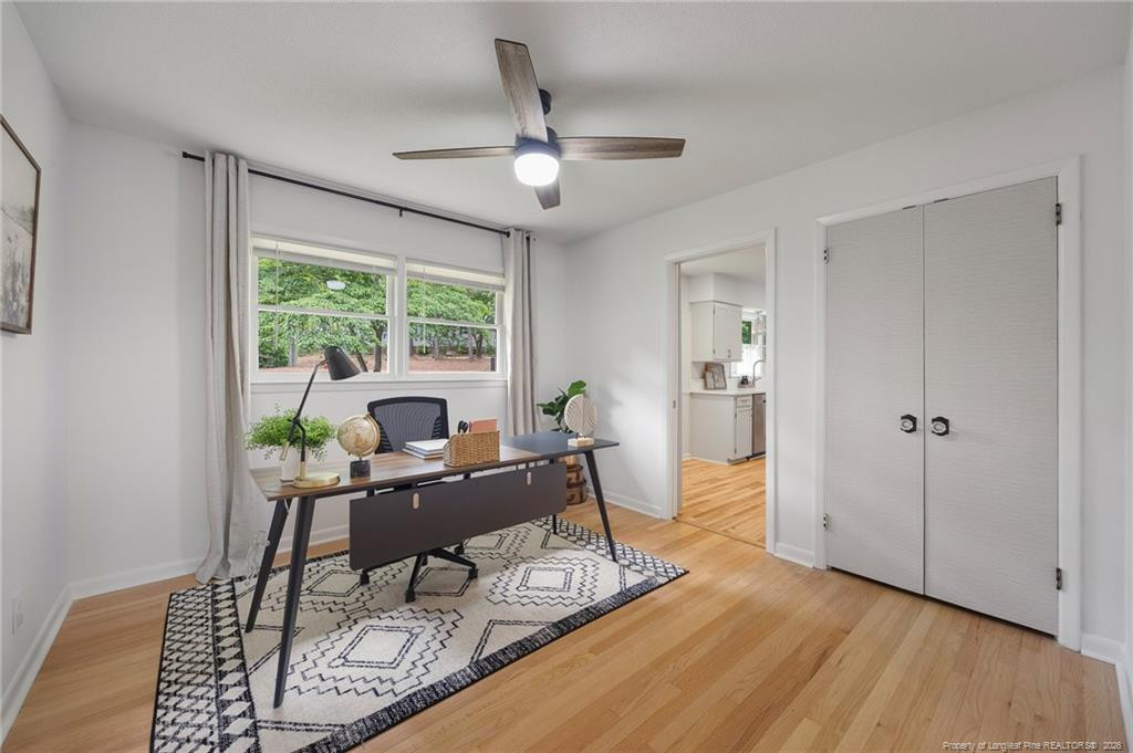 Furnished home office with a desk beneath windows overlooking trees and hardwood floors.