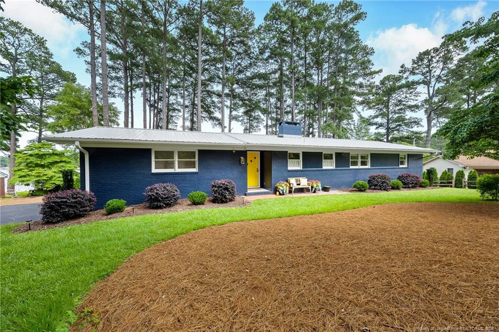 Blue ranch-style home with a bright yellow front door, landscaped lawn, and tall pines behind it.