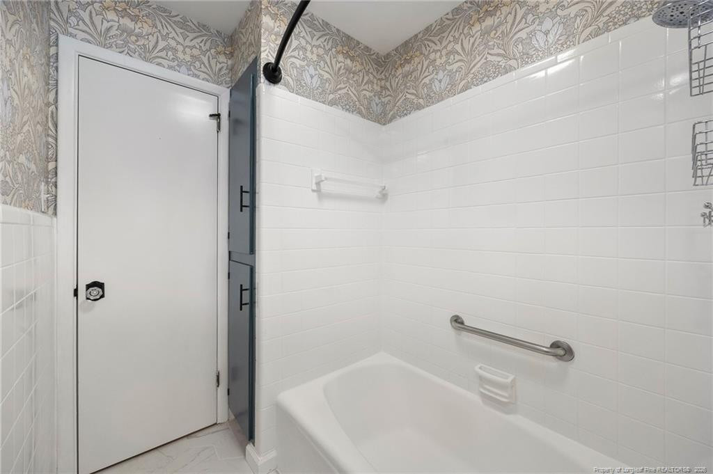 Bathroom with a white tiled tub and shower, grab bar, and patterned wallpaper.