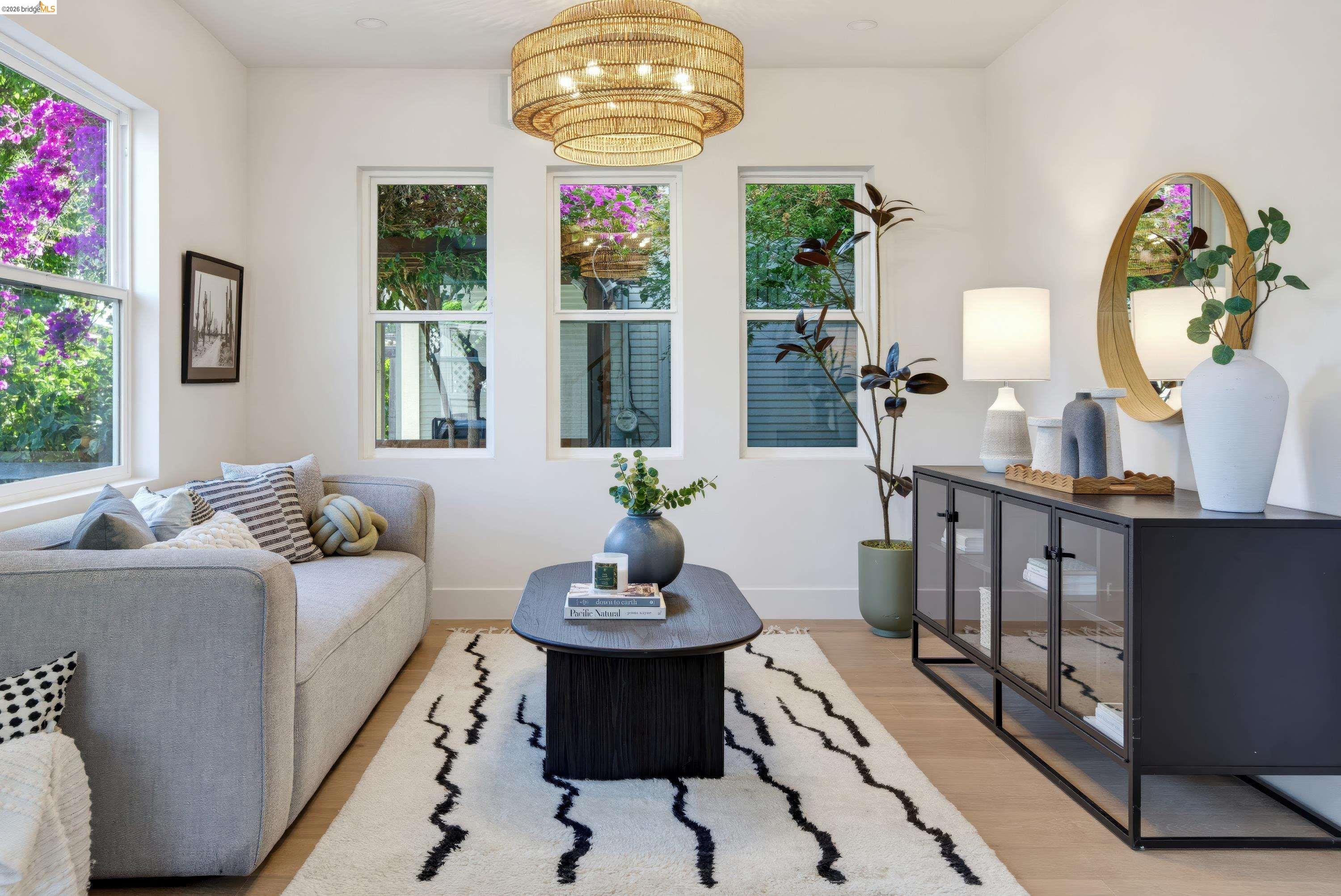 Staged living room with a gray sofa, patterned rug, and gold chandelier beneath windows overlooking greenery.