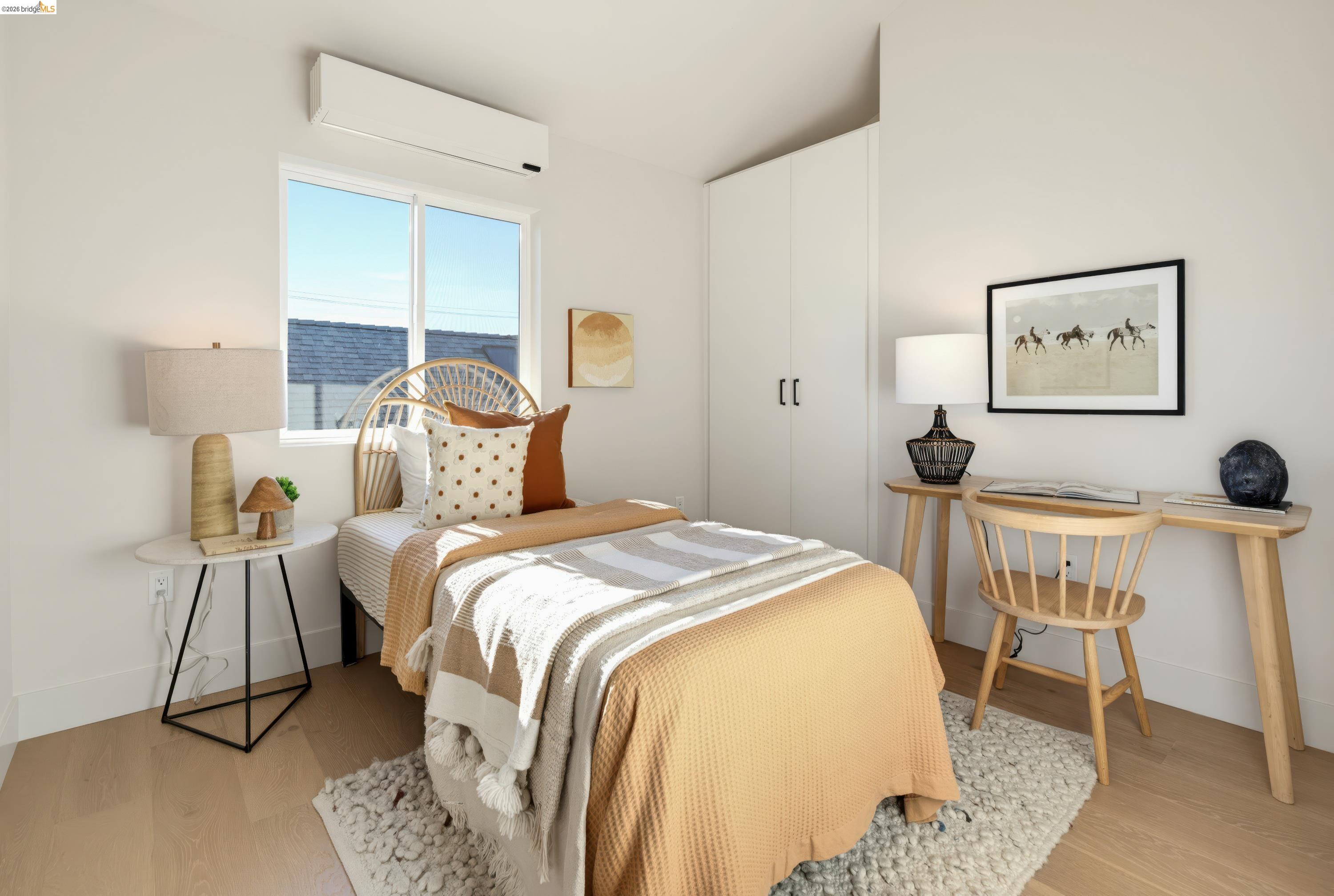 Bright staged bedroom with a tan-and-cream bed, large window, built-in closet, and small desk.