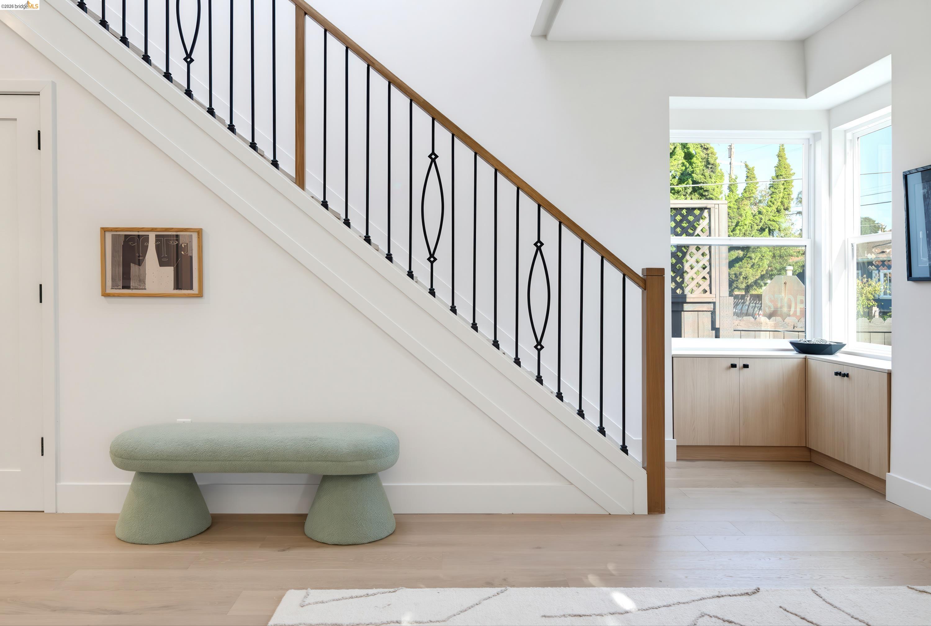 Bright entryway with a black iron staircase, sage green bench, and light wood floors.