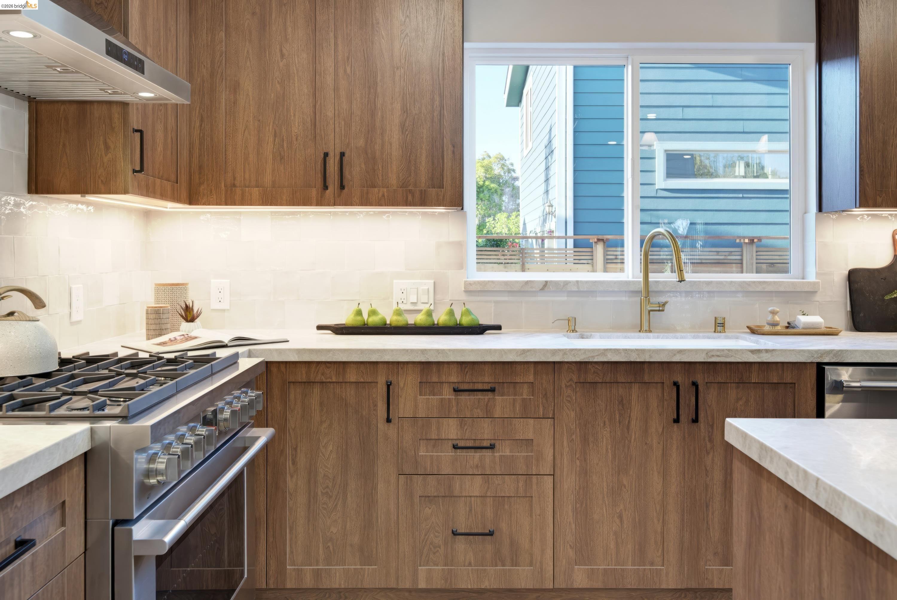 Modern kitchen with warm wood cabinets, stainless steel range, and a large window above the sink.