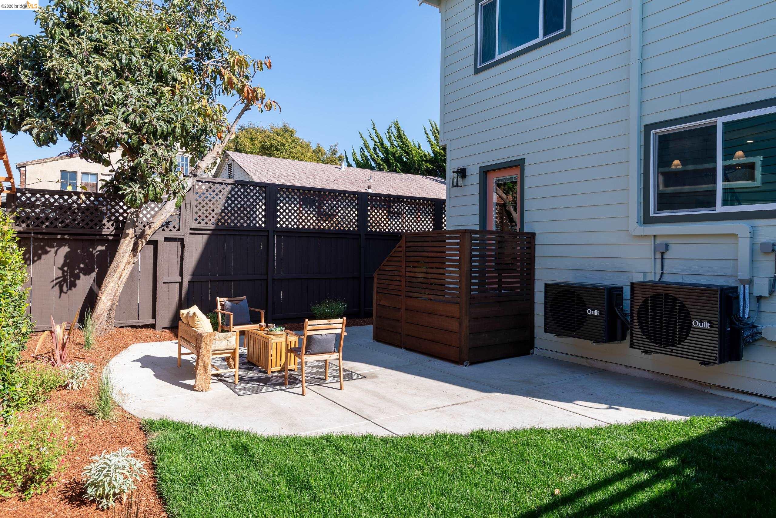 Backyard patio with outdoor seating, a tall privacy fence, mature tree, and green lawn.