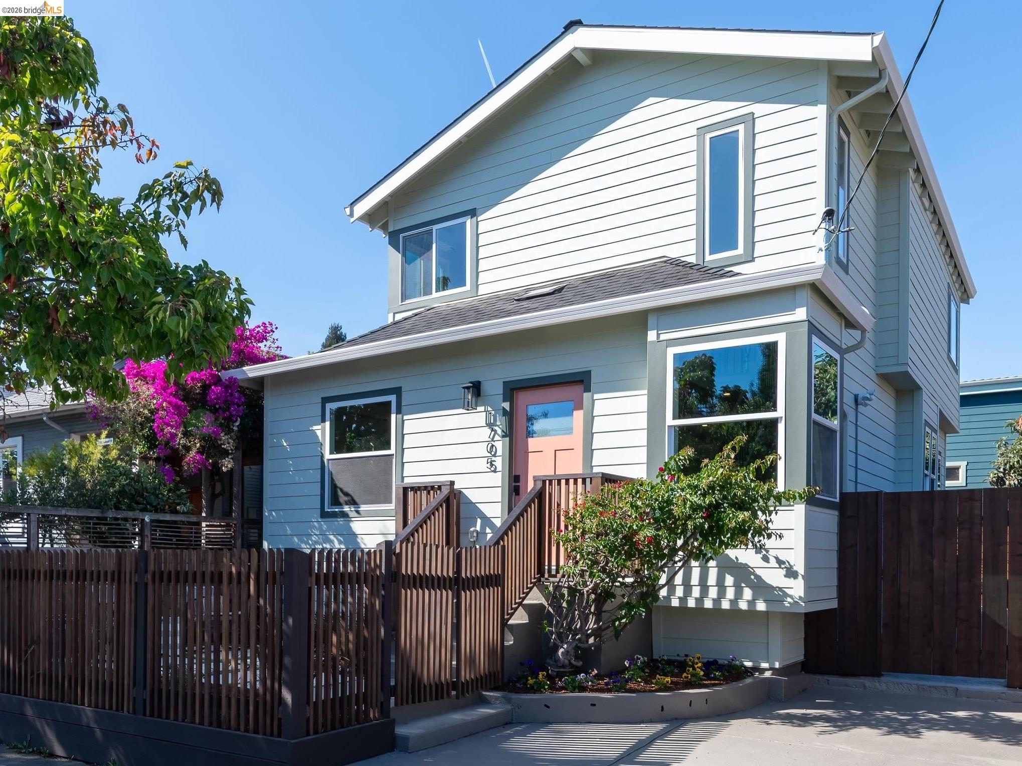 Two-story blue-gray home with a pink front door, wooden stairs, and flowering bougainvillea.