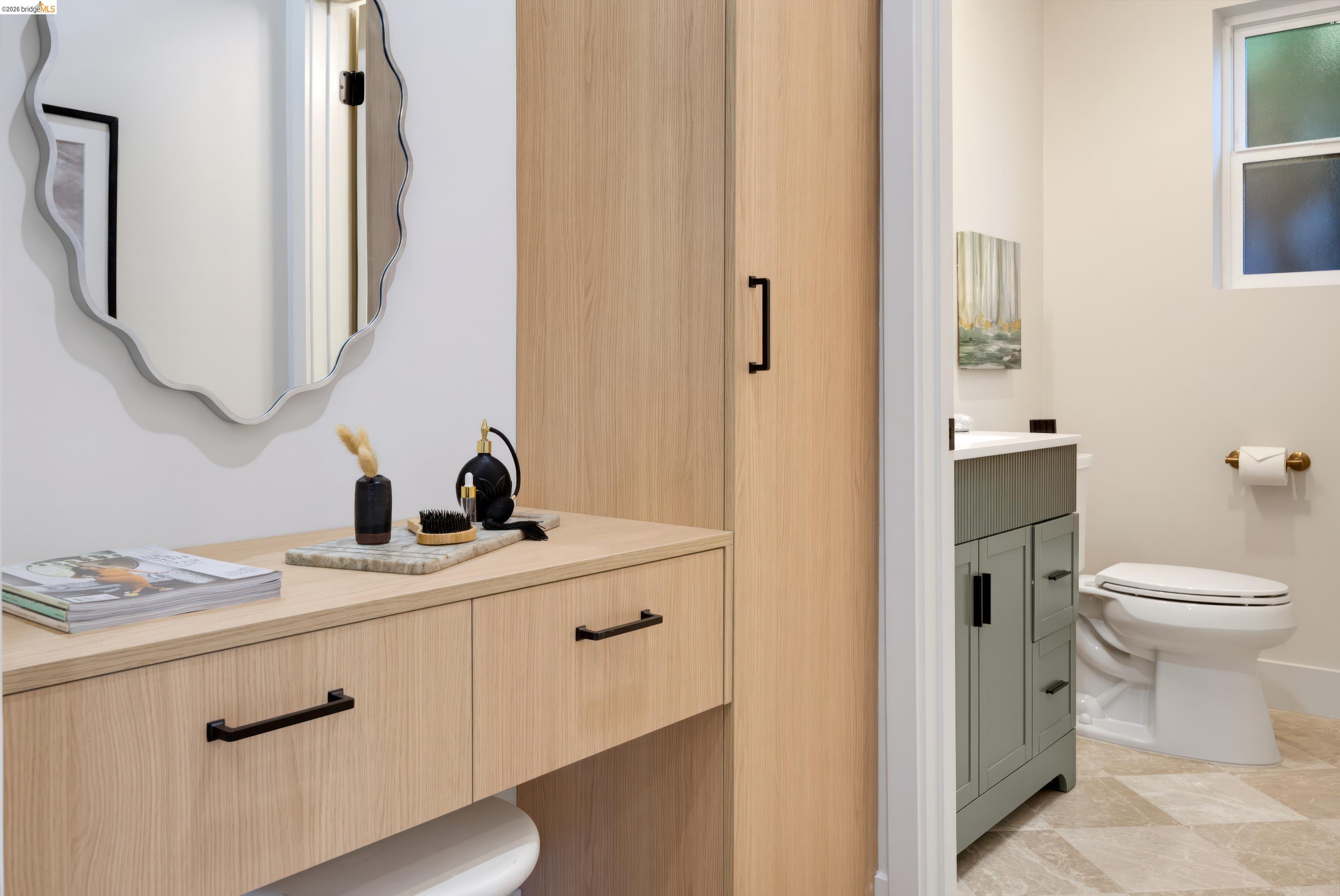 Bathroom with a light oak vanity, wavy-edged mirror, sage-green sink cabinet, and marble-look tile floor.
