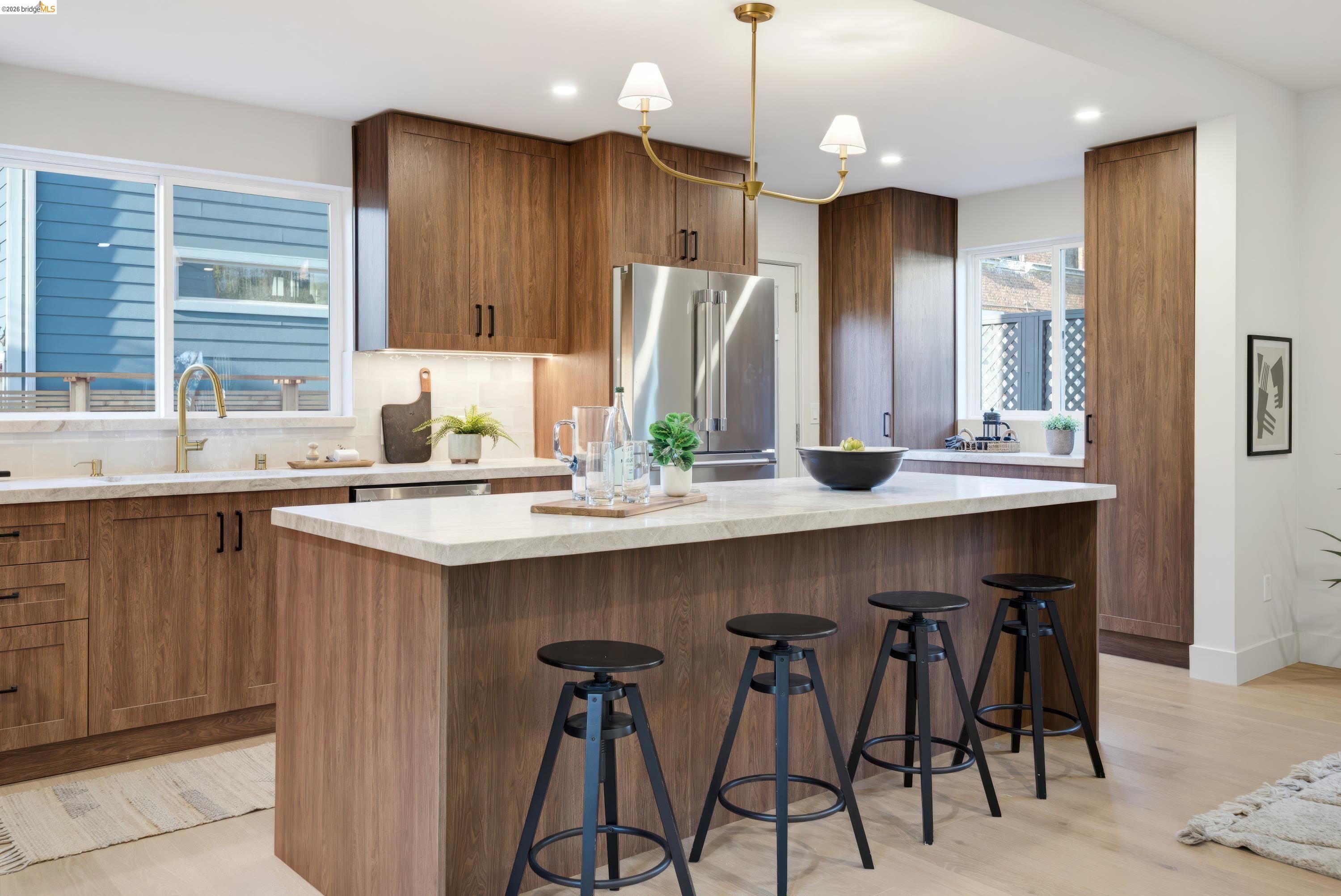 Kitchen with walnut cabinets, a large stone island, four black stools, and a stainless steel refrigerator.
