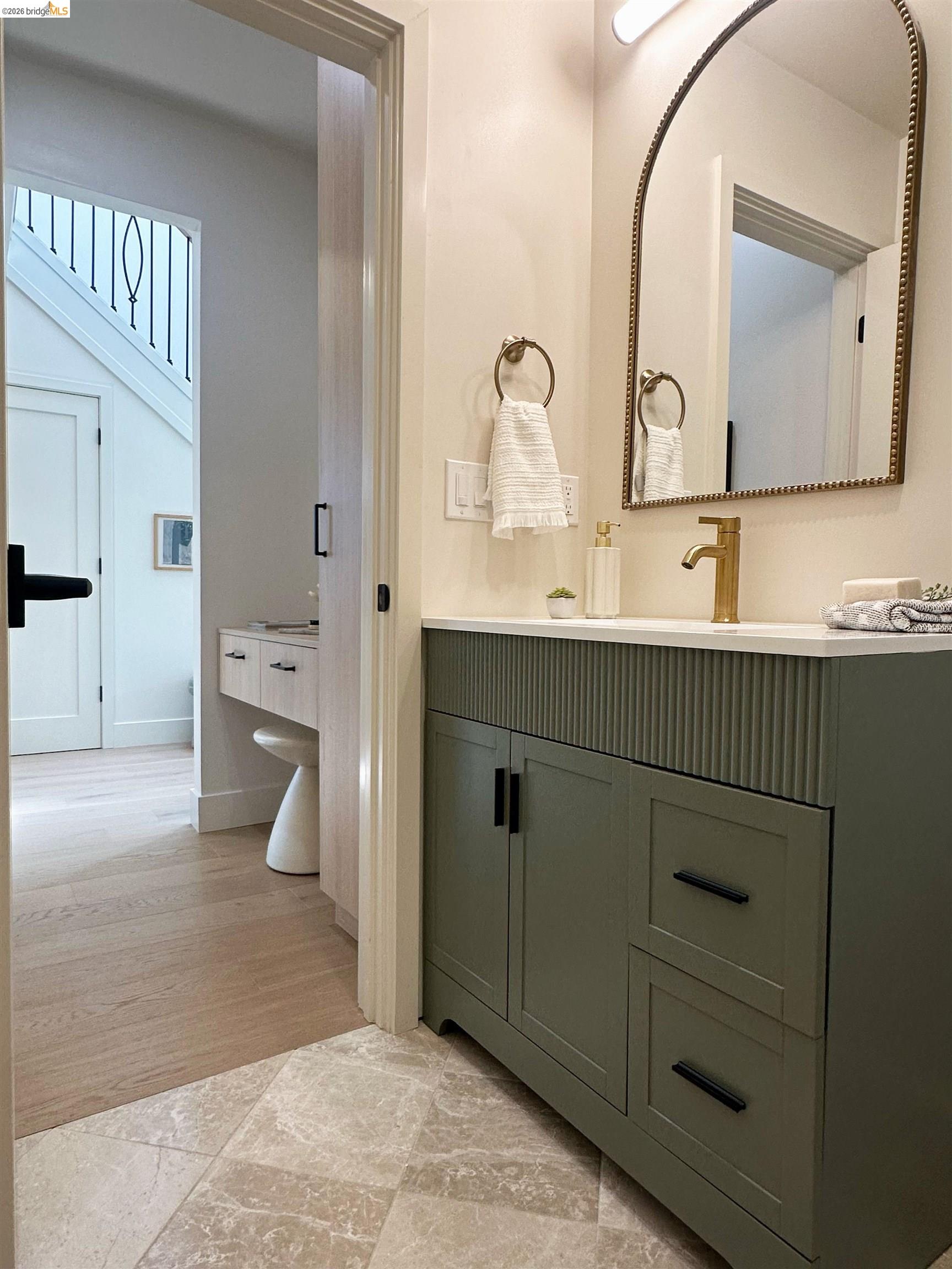 Bathroom with a fluted green vanity, brass faucet, and arched mirror beside an open hallway doorway.