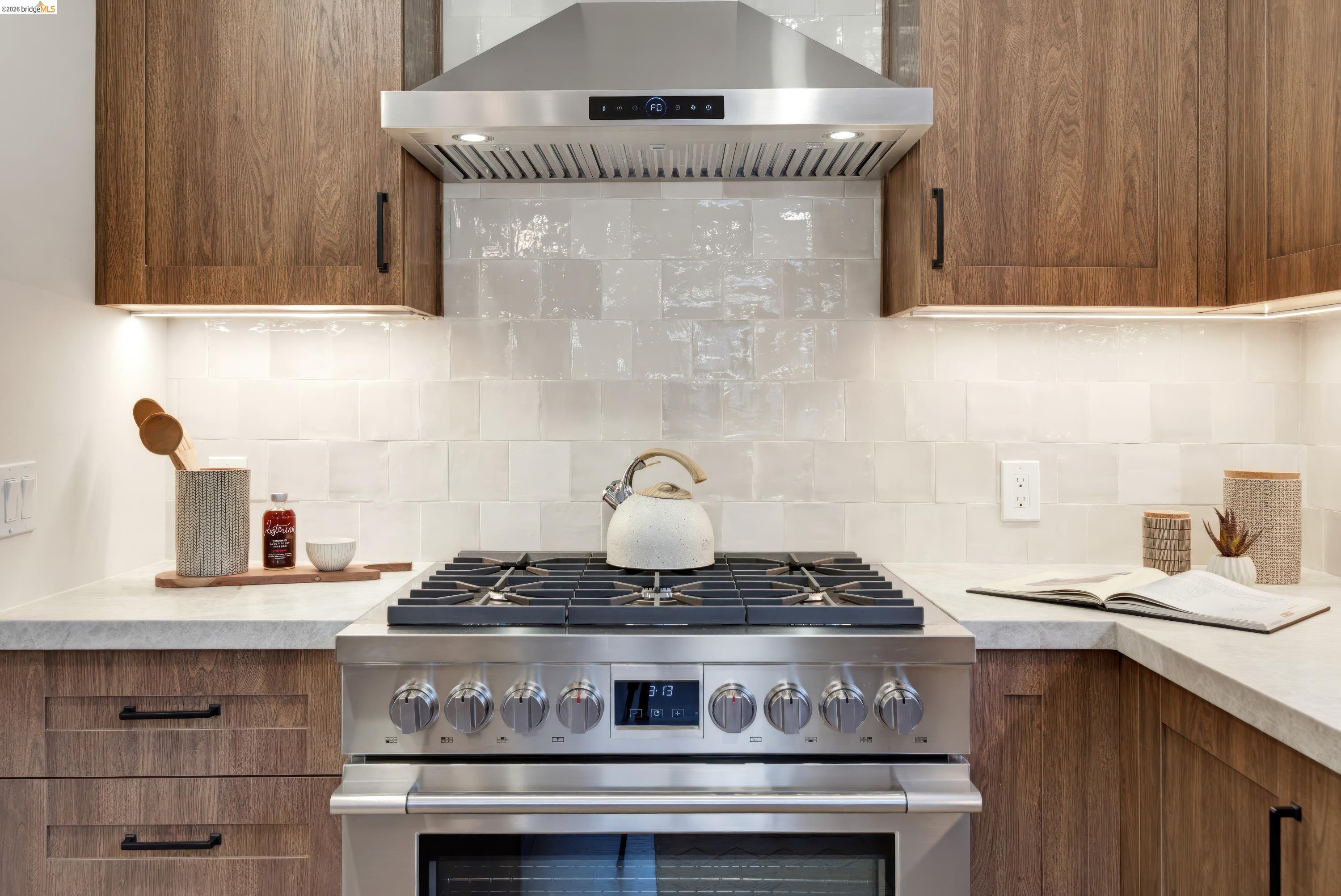 Kitchen range with stainless steel gas cooktop and hood, walnut cabinets, and glossy white tile backsplash.