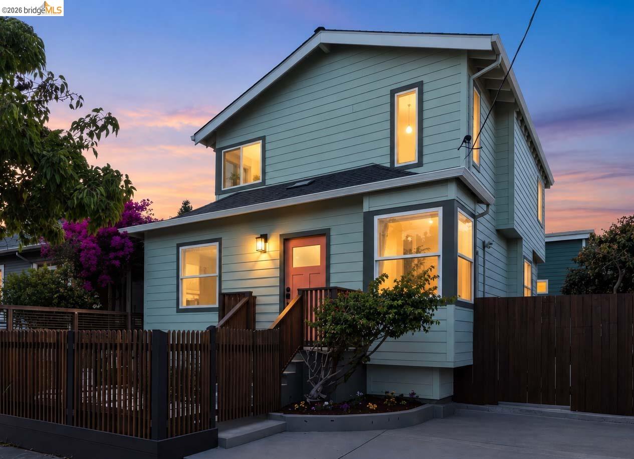 Two-story blue-gray house with a red front door and warmly lit windows behind a wooden fence at dusk.