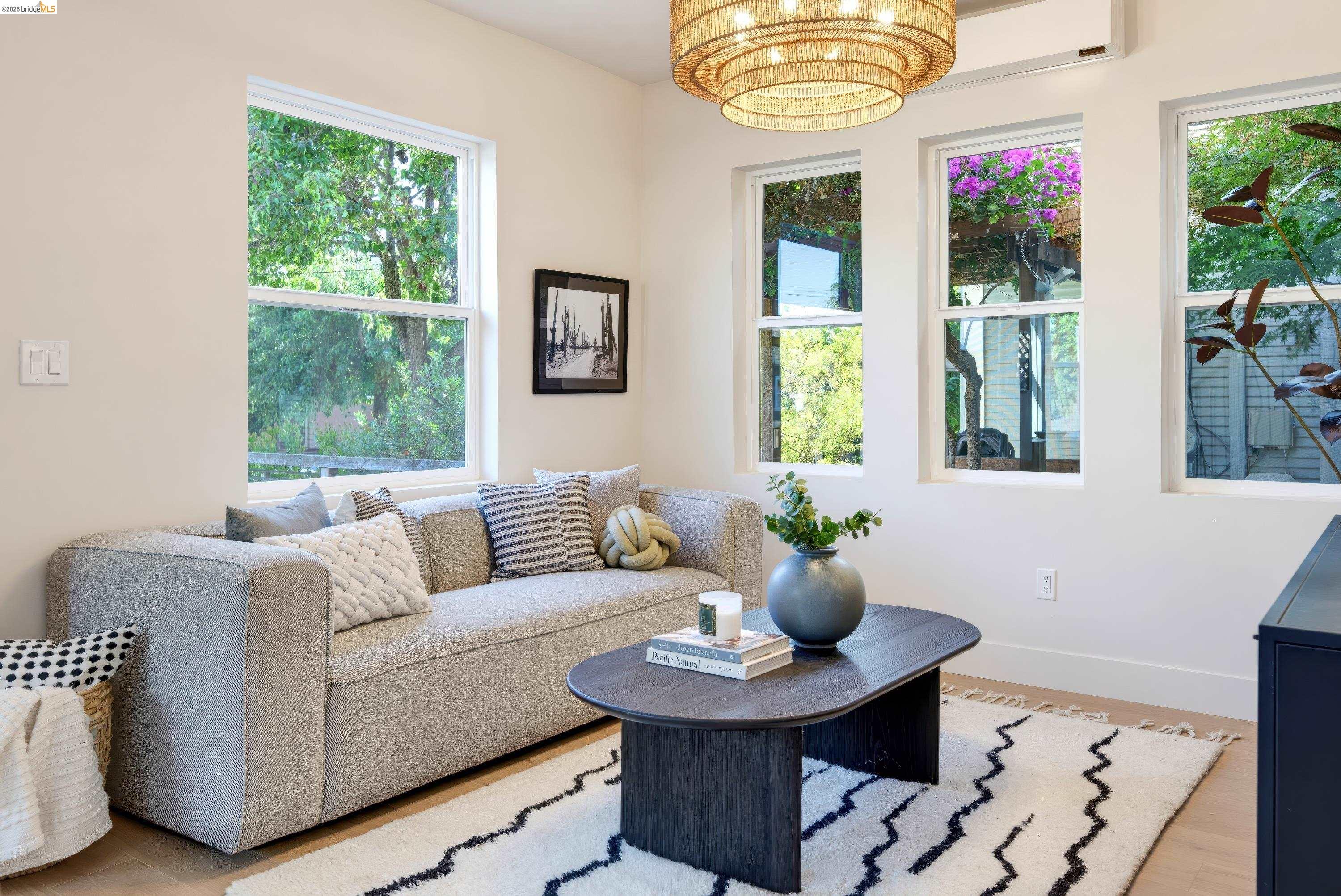 Staged living room with a gray sofa, patterned rug, and tall windows overlooking greenery and purple flowers.