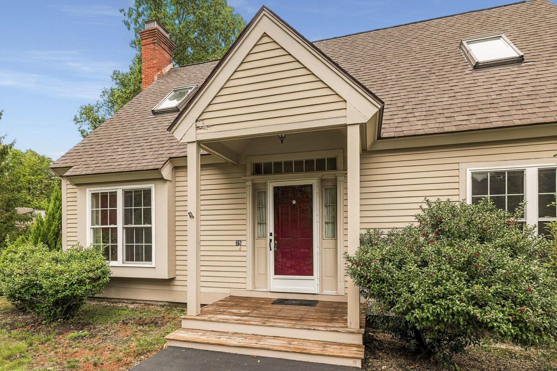 Beige-sided house with a red front door beneath a covered porch, surrounded by shrubs.