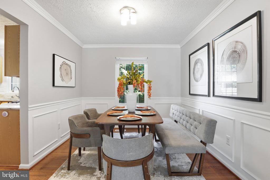 Dining room with a set table, gray bench, white wainscoting, and flowers by the window.