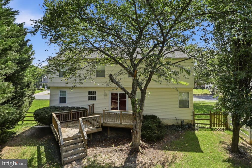 Rear of a two-story house with a raised wooden deck, stairs, and mature trees.