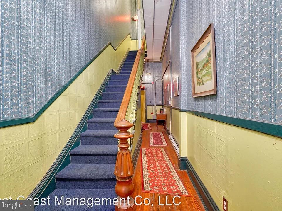 Hallway with a blue-carpeted staircase, polished wood banister, patterned wallpaper, and red runner rugs.