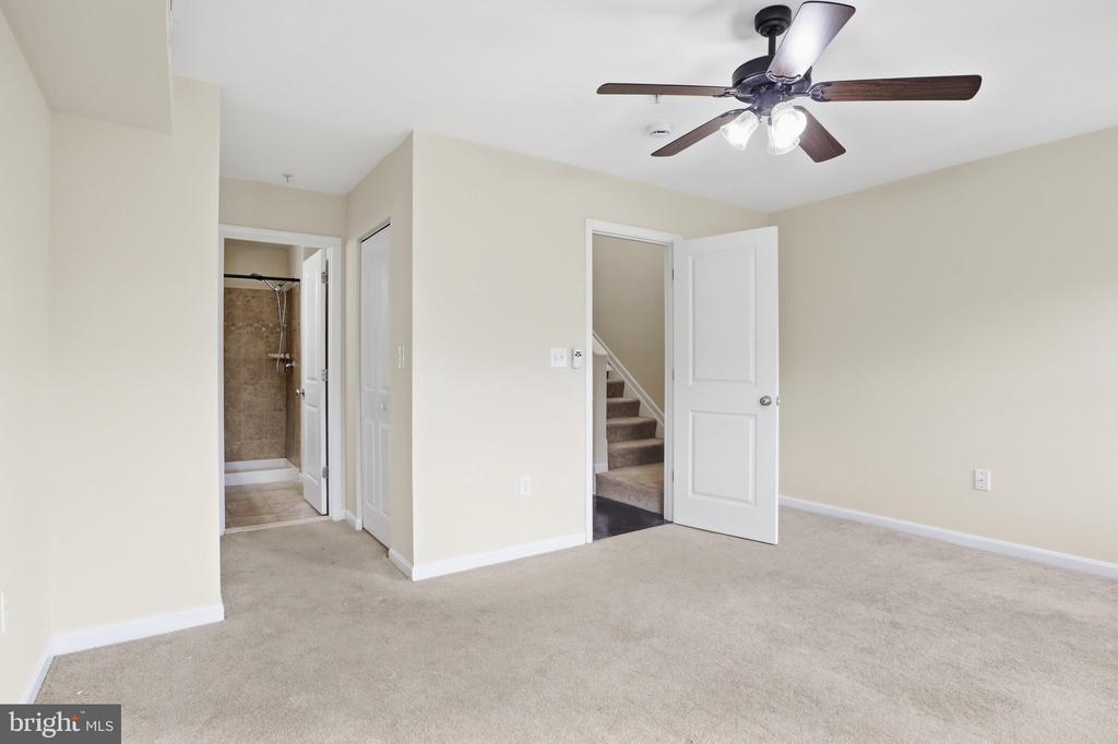 Empty carpeted bedroom with a ceiling fan and open doorways to a shower and stairs.