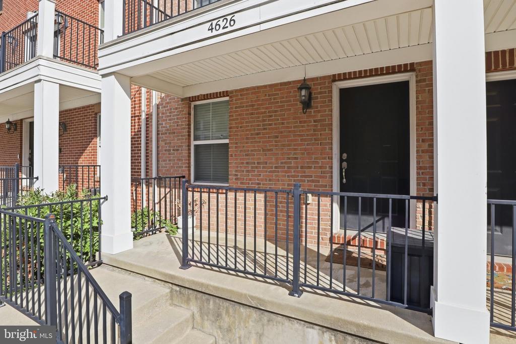 Brick home entrance with covered porch, white columns, black railings, and concrete steps.