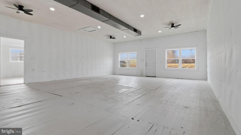 Vacant room with a worn plank floor, high ceiling, black fans, and windows overlooking open fields.