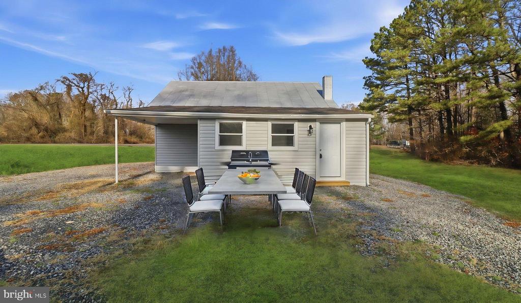 Gray-sided house with a covered patio, outdoor dining set, and barbecue beside a gravel drive.