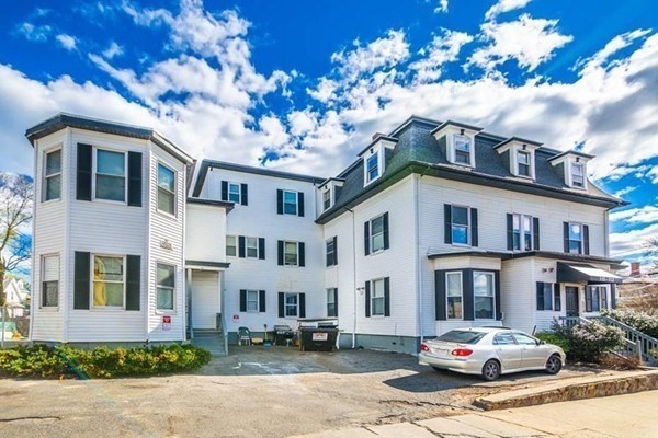 White multi-unit building with dormer windows and a silver car parked in front.