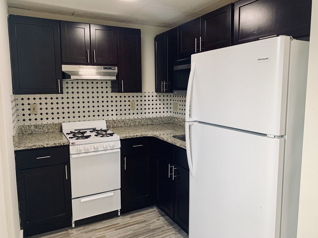 Compact kitchen with dark cabinets, diamond-pattern backsplash, granite counters, and white appliances.