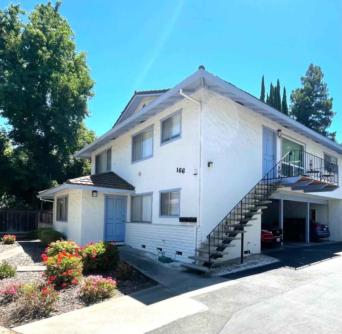 White two-story residence with blue doors, an exterior staircase, carport, and flower beds.