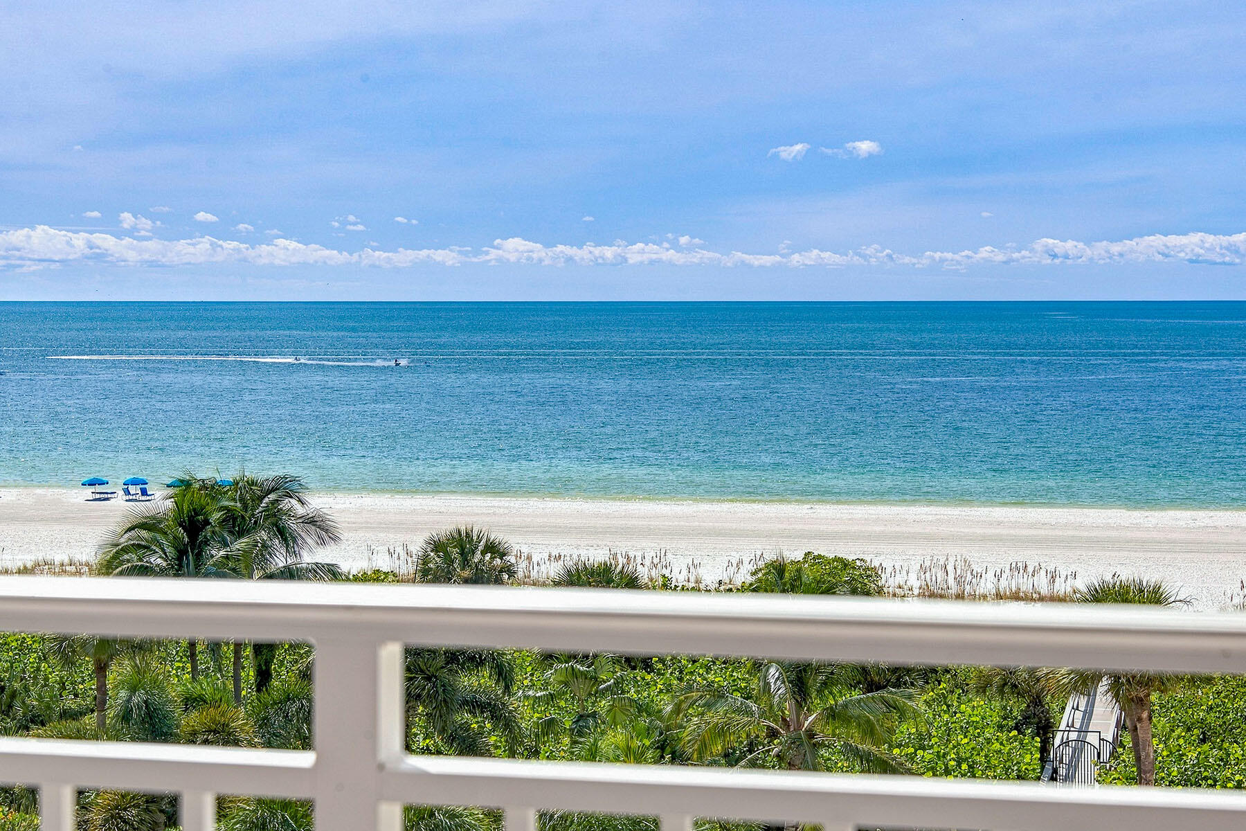 Ocean and white-sand beach seen over a balcony railing, with palm trees and blue beach umbrellas.