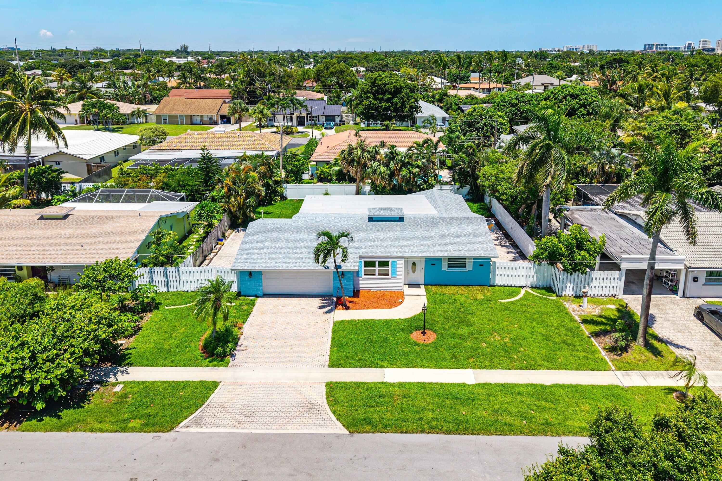 Aerial view of a turquoise-and-white house with a gray roof, paver driveway, and palm-lined yard.
