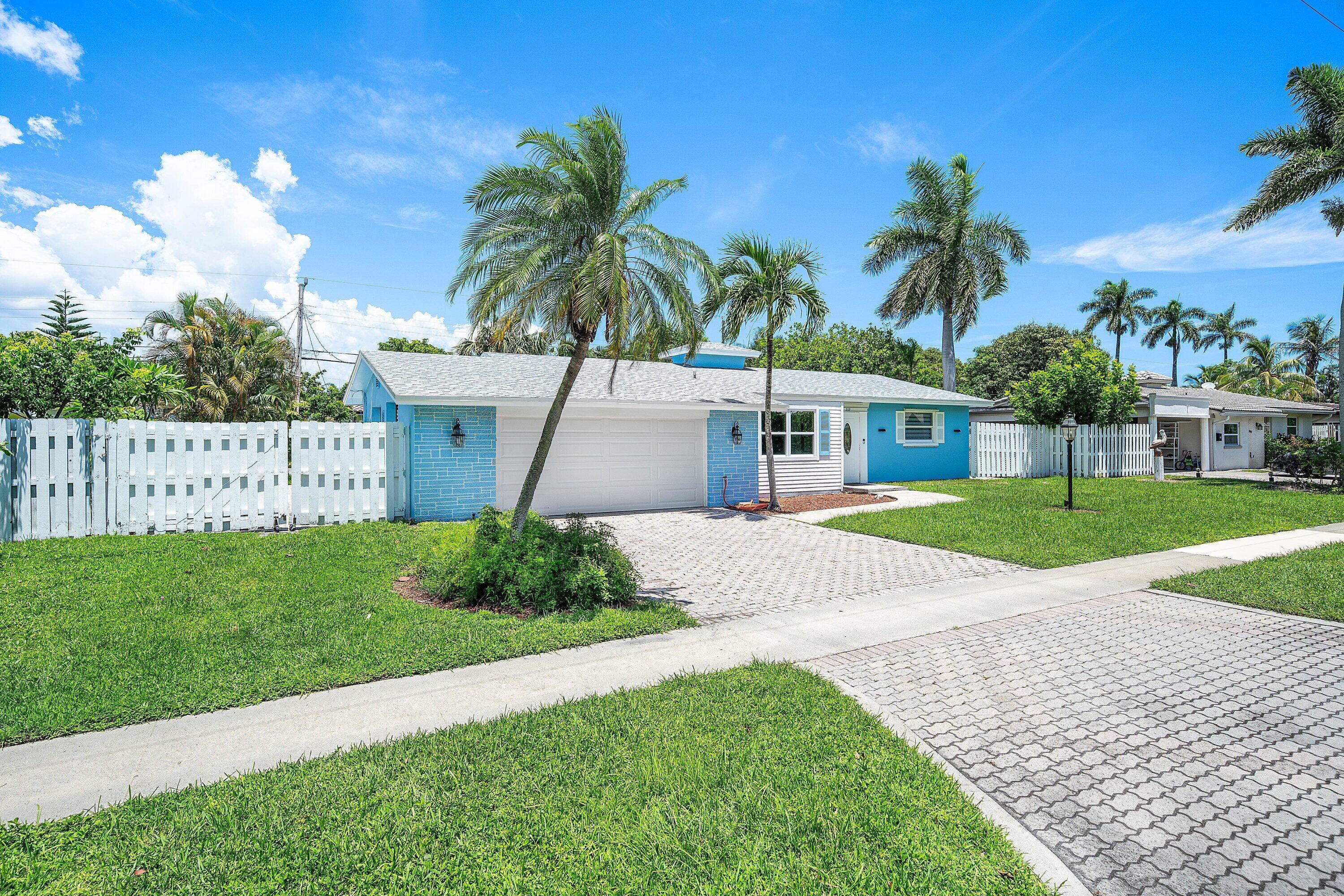 Blue single-story house with a white garage, picket fence, palm trees, and paver driveway.
