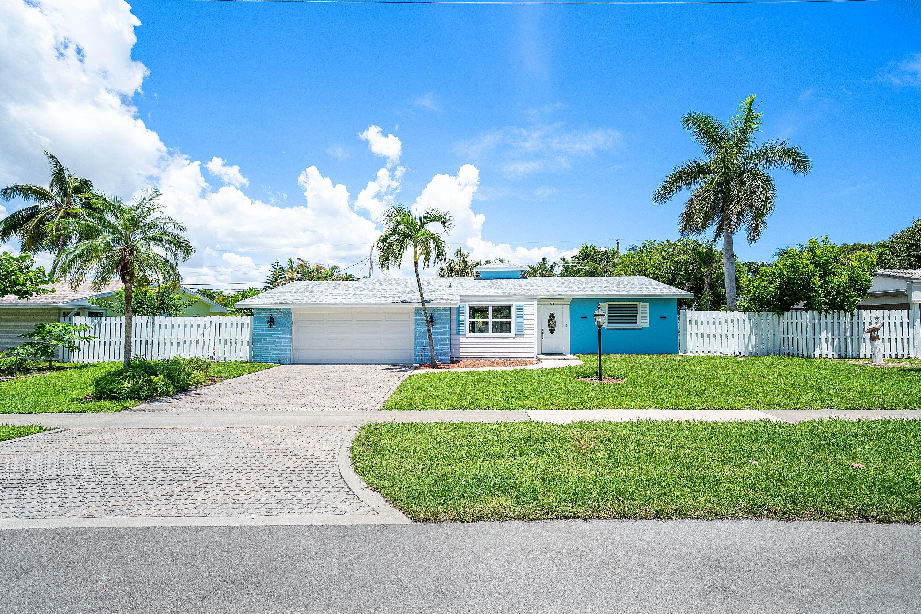 Blue-and-white single-story home with a paved driveway, white fencing, and palm trees.