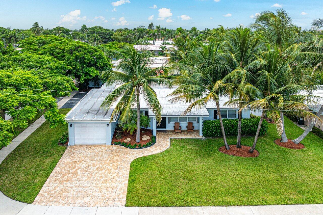 Aerial view of a light blue home with a curved paver driveway, palm trees, and a wide lawn.