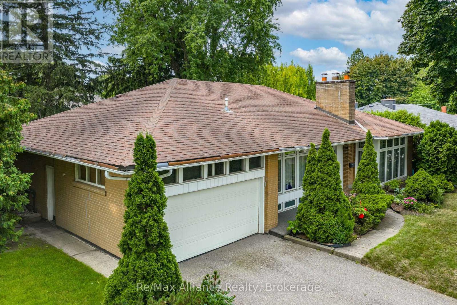 Single-storey brick house with a broad shingle roof, double garage, and mature trees and shrubs.