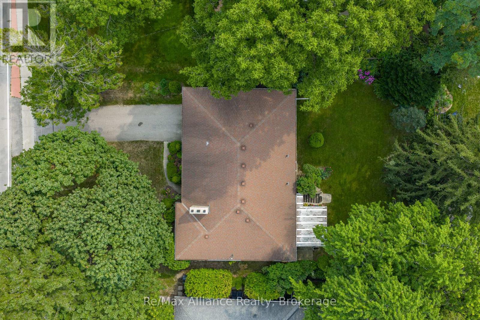 Aerial view of a brown-roofed house surrounded by mature trees, lawn, and a driveway.