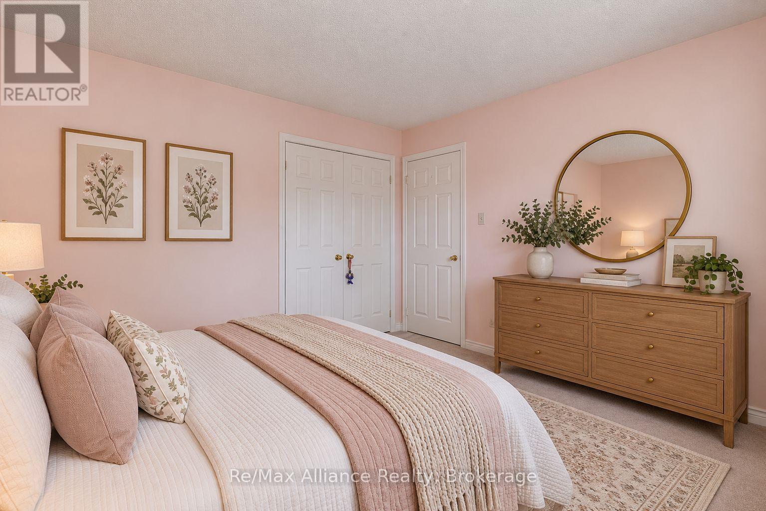 Staged bedroom with blush bedding, pink walls, botanical prints, and a wooden dresser beneath a round mirror.
