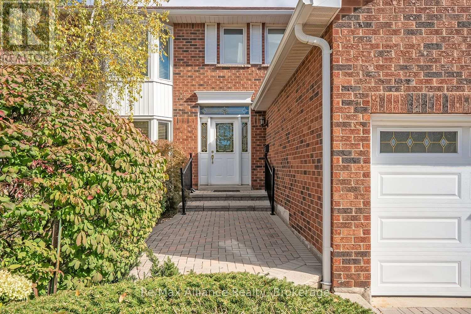 Red-brick home entrance with decorative glass door, paved walkway, shrubs, and a white garage door.