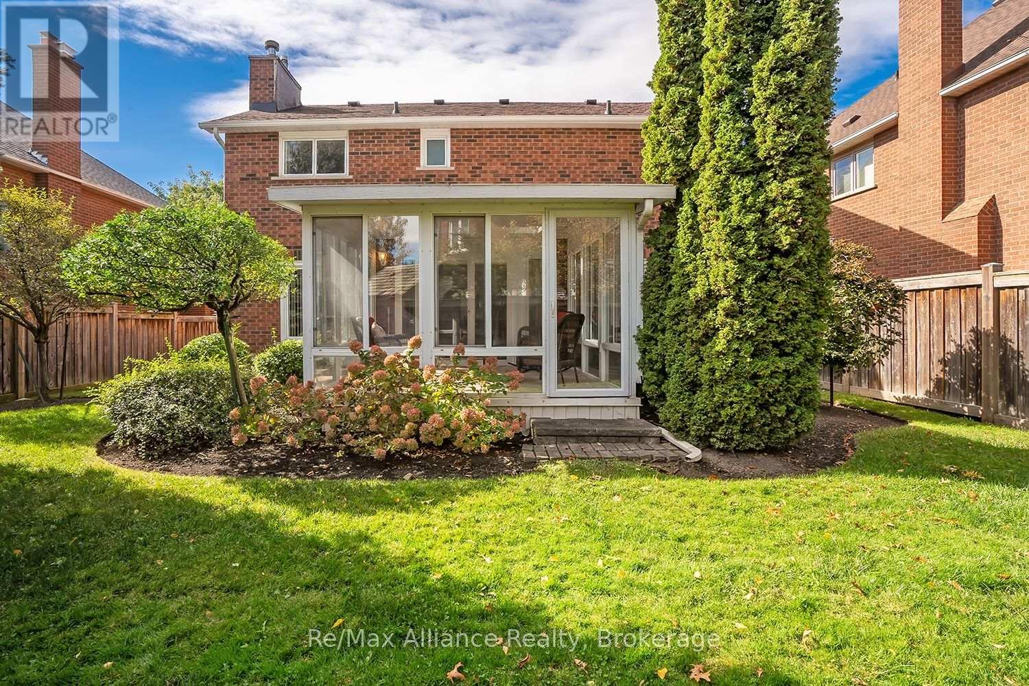 Brick home’s rear sunroom overlooks a fenced lawn with mature landscaping and a small paved patio.