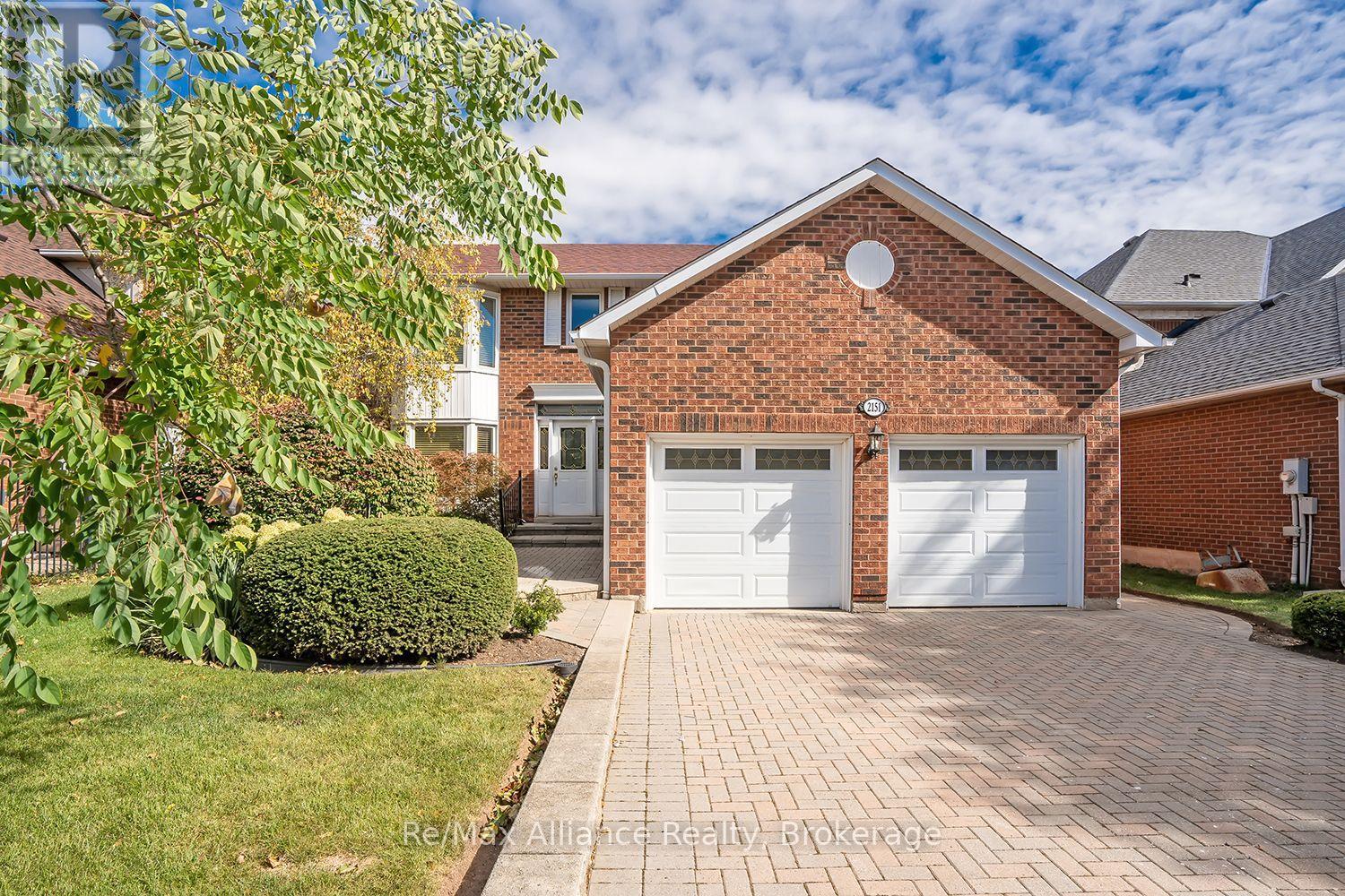 Brick home with a double garage, broad paver driveway, and leafy landscaping.