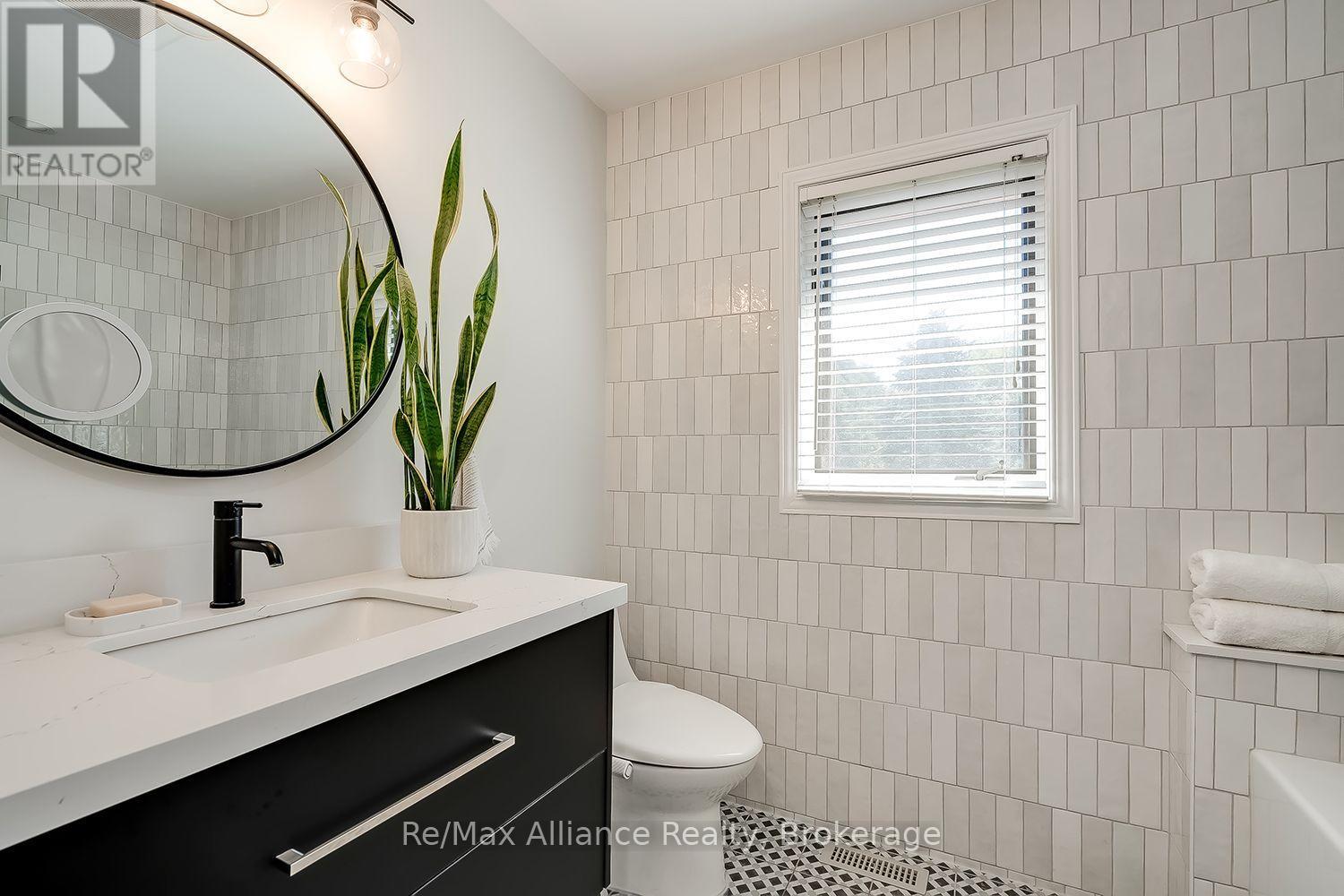 Bathroom with white tiled walls, black vanity, oval mirror, and window with blinds.