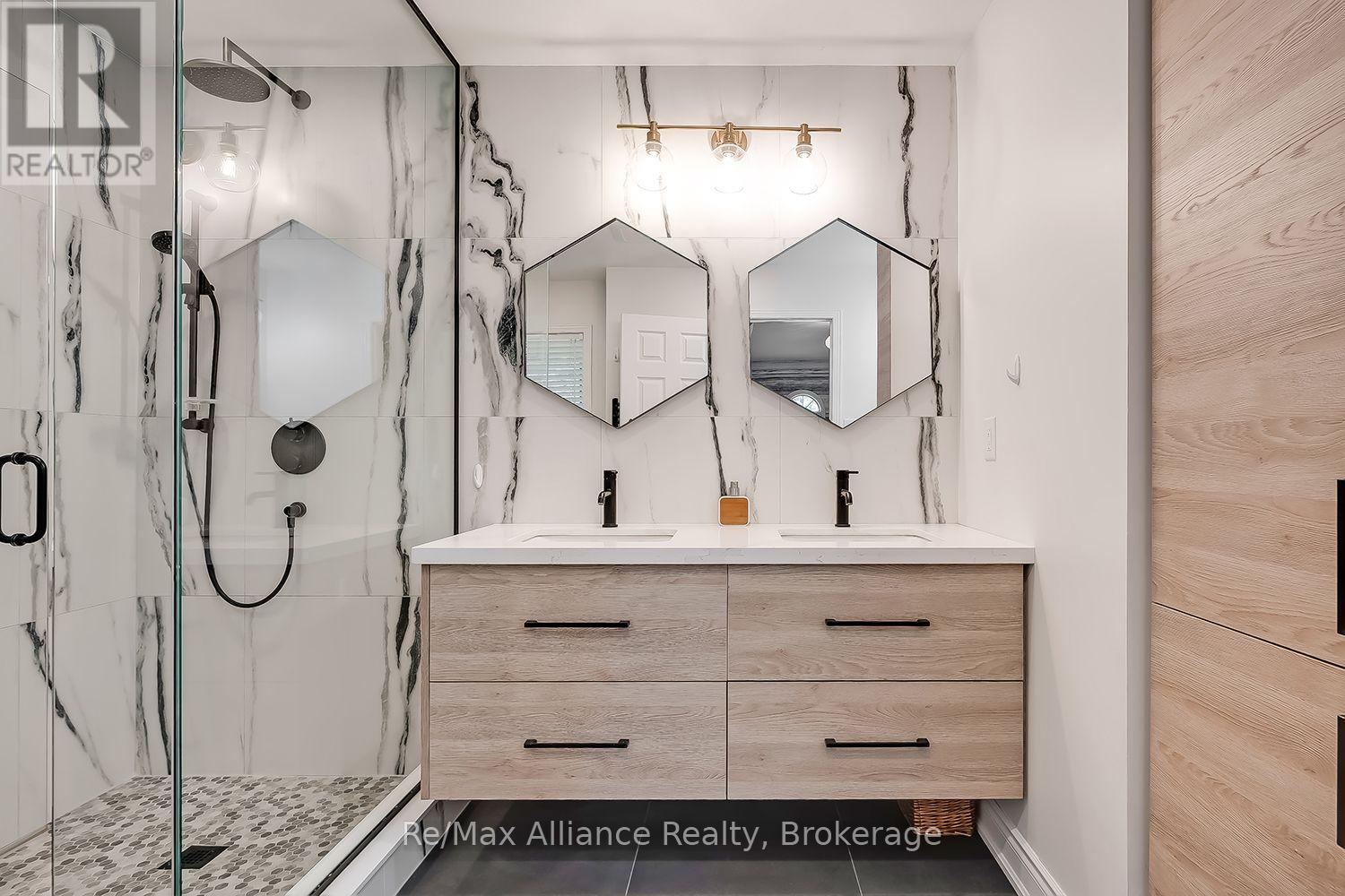 Bathroom with a glass walk-in shower, pebble-tile floor, and light wood double vanity beneath geometric mirrors.