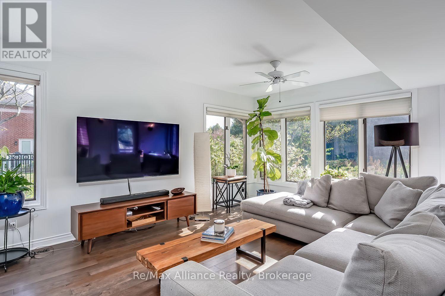 Furnished living room with a white sectional, wall-mounted television, and broad windows overlooking greenery.