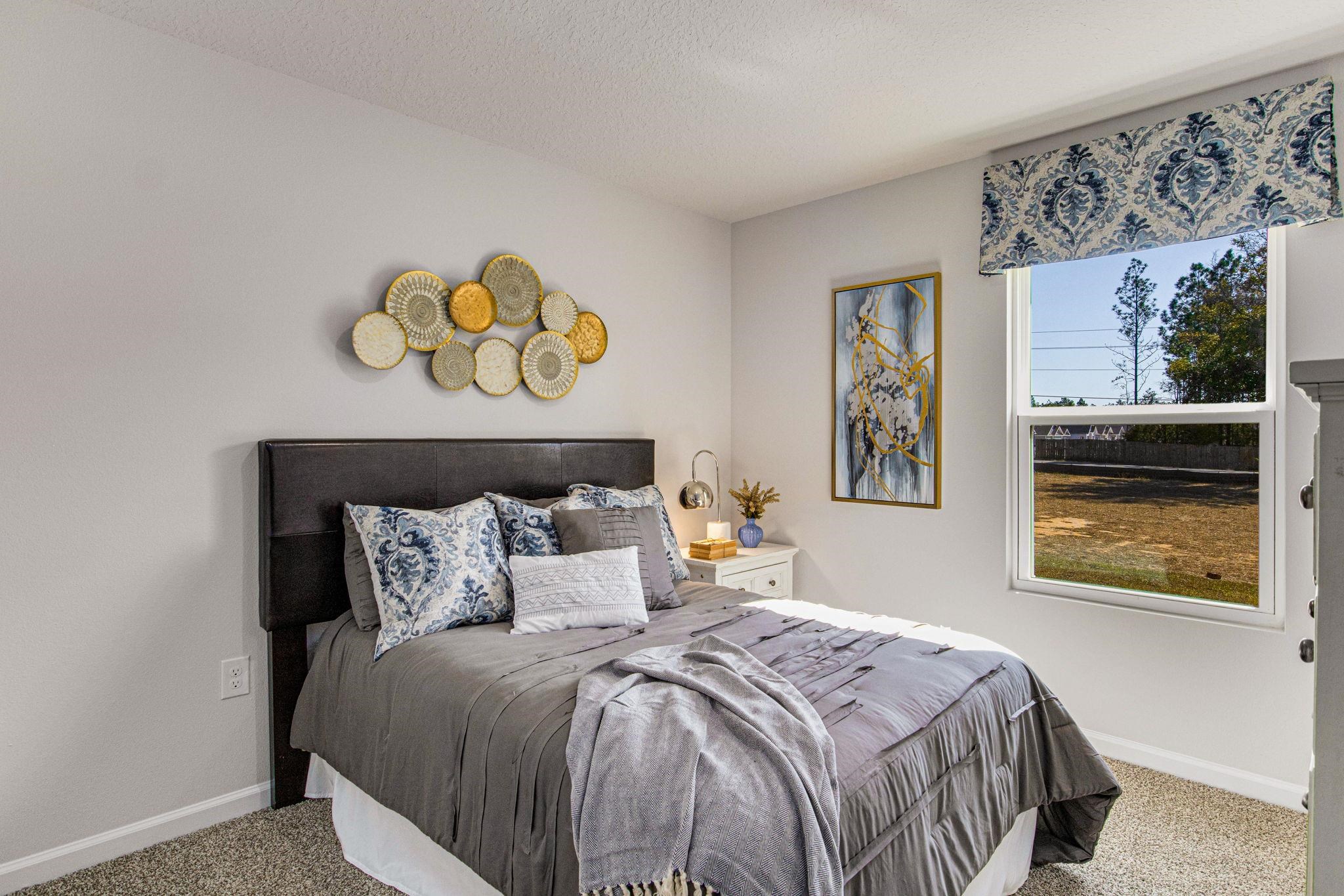 Staged bedroom with a gray bed, patterned pillows, circular wall decor, and a large yard-facing window.