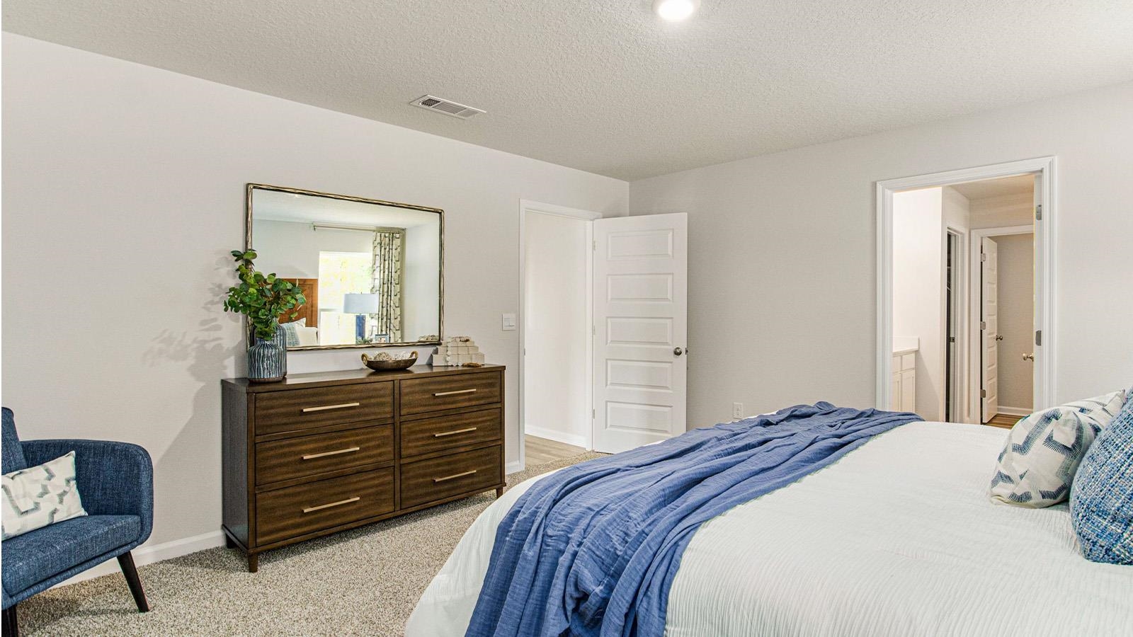 Staged bedroom with a white bed, blue throw, dark dresser, and adjoining doorway.