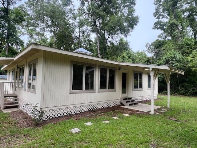 Single-story light-sided house with a long covered porch and lattice skirting, surrounded by mature trees.