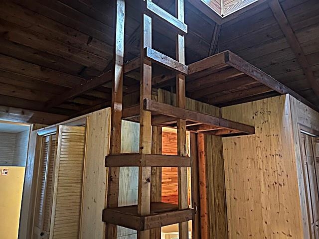 Wood-paneled interior with exposed beams, plywood partitions, and a tall ladder-like structure leading to an elevated platform.