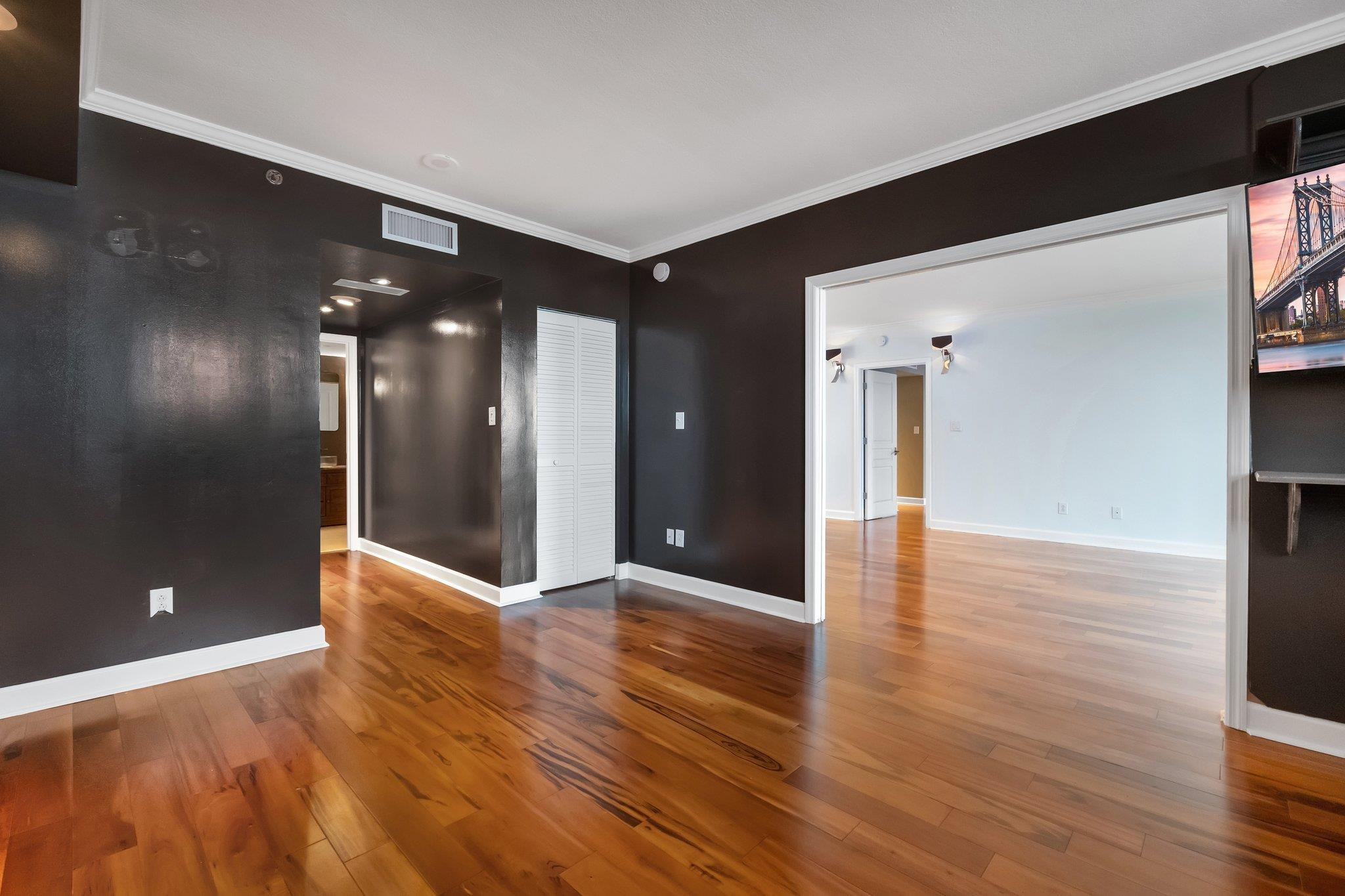Empty condo living area with glossy hardwood floors and dark brown walls opening into a bright adjoining room.
