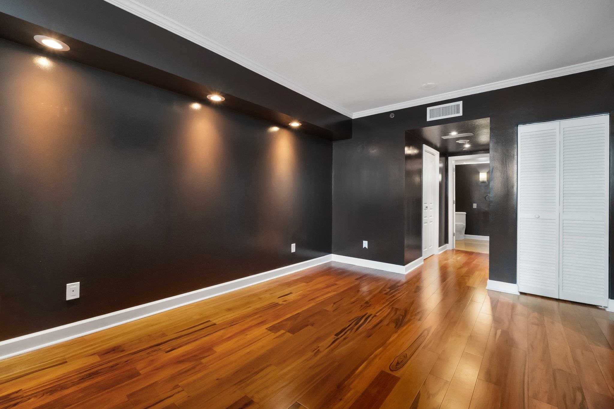Empty condo room with dark walls, hardwood floors, and a louvered closet beside a bathroom doorway.