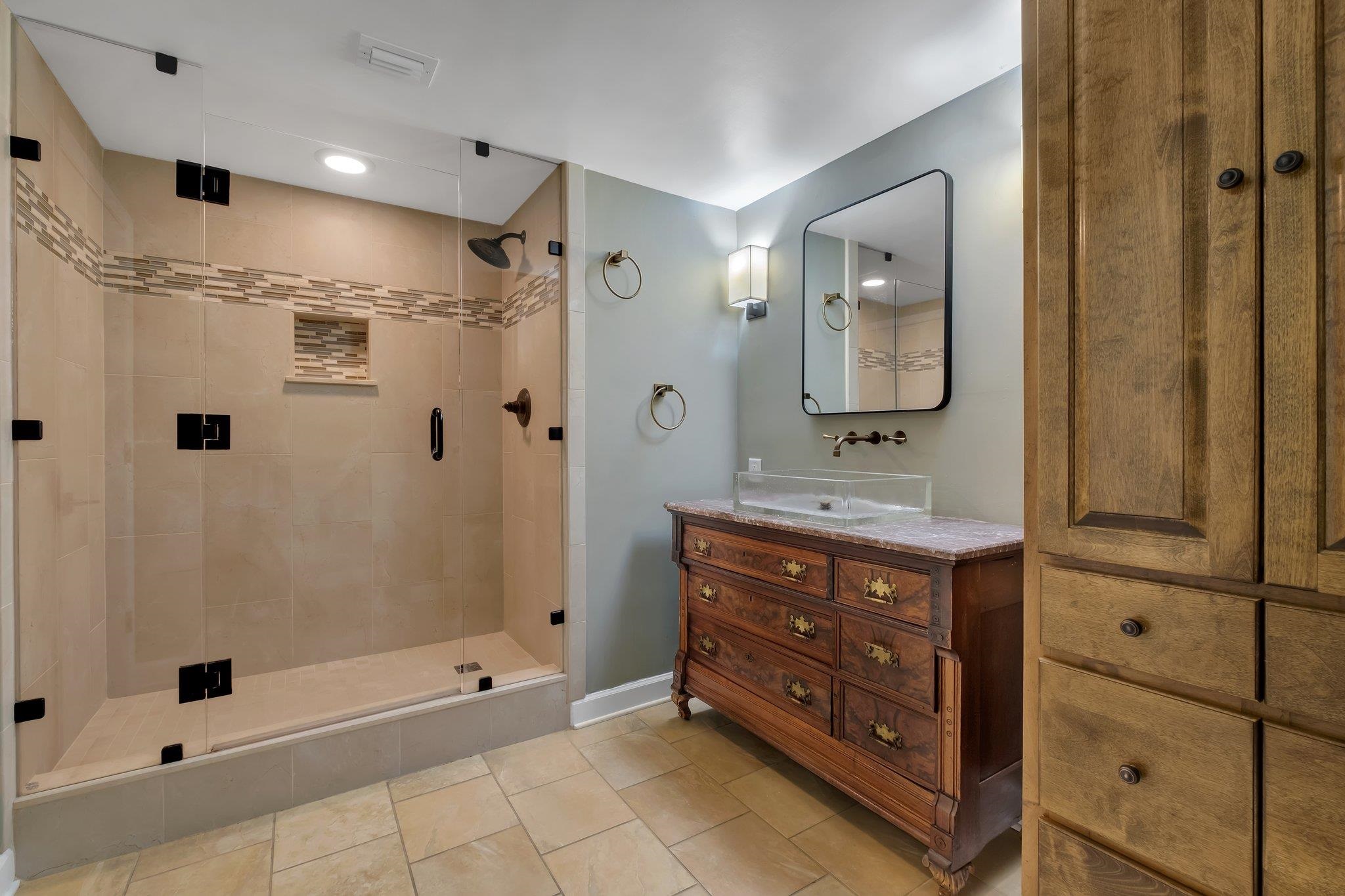 Bathroom with glass-enclosed tiled shower, clear vessel sink, ornate wooden vanity, and tall linen cabinet.