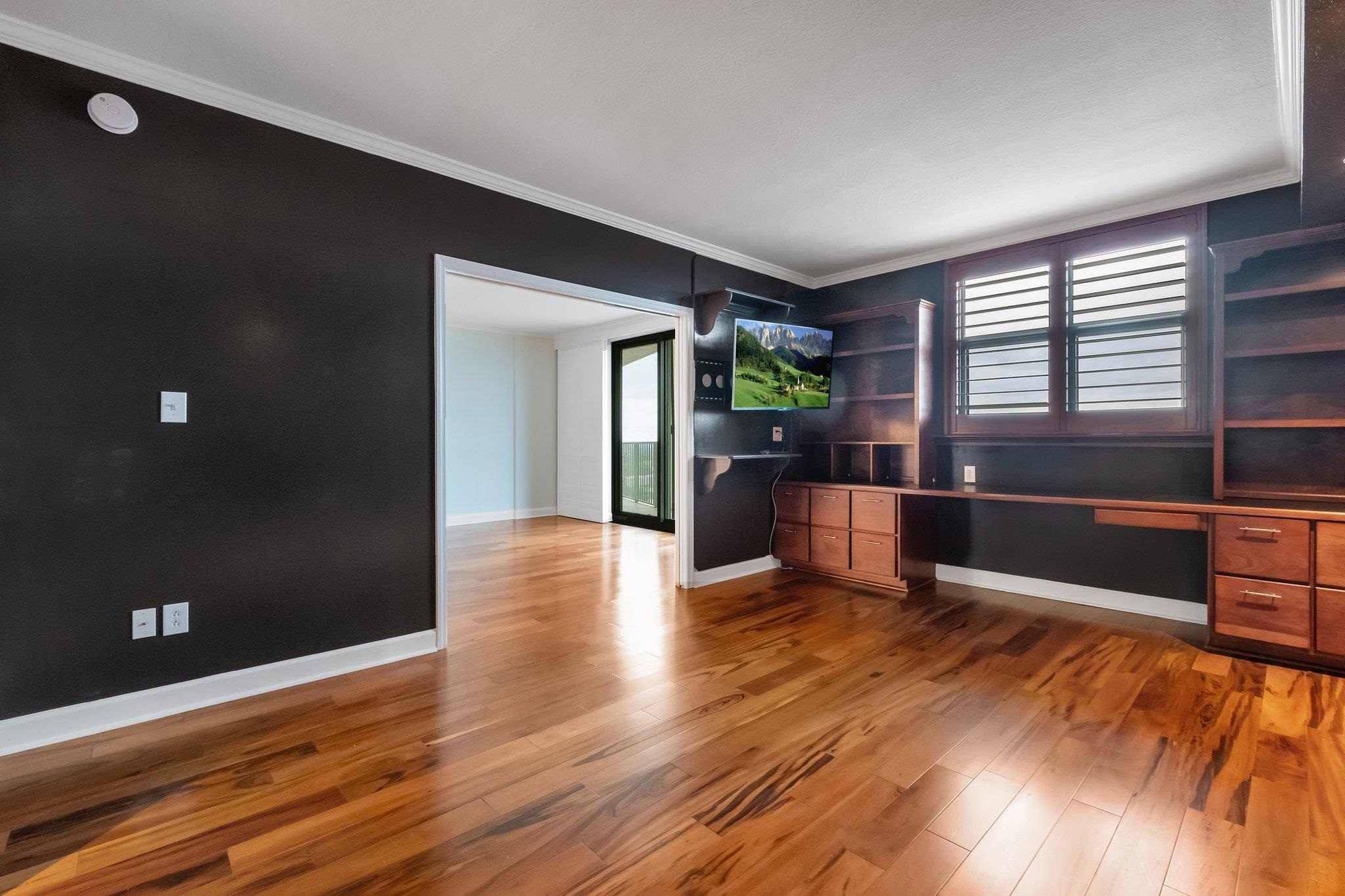 Empty condo office with built-in wood desk and shelving, plantation shutters, and hardwood floors.