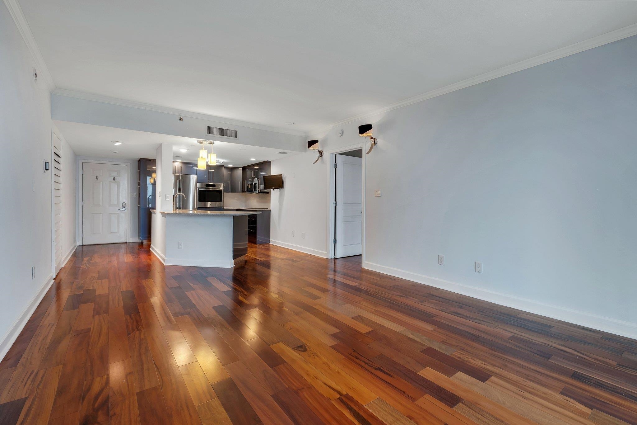Open condo living area with glossy hardwood floors and an adjoining kitchen with dark cabinets and stainless steel appliances.